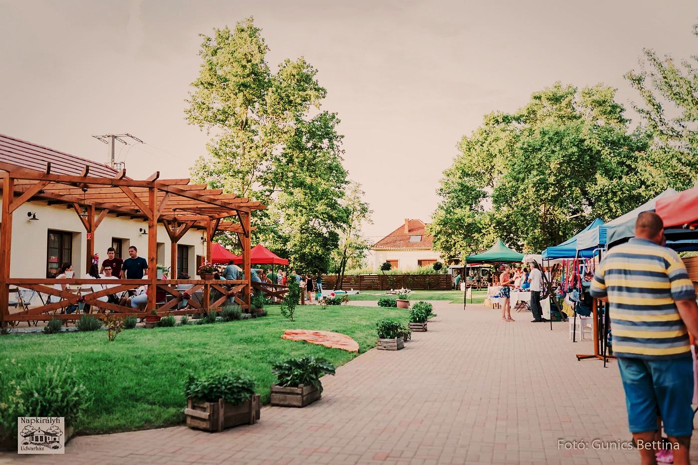 Vásári standok sora az udvarház bejárati útján, a pergola alatt ülő vendégekkel